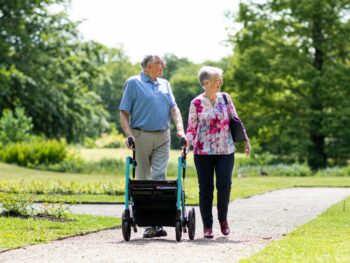 A man and woman walk down a path. The man is using a four-wheeled Rollator.
