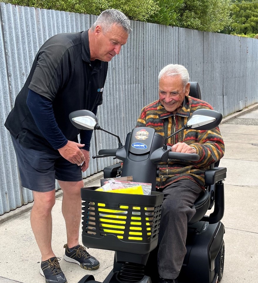 Mobility Centre staff, Gary, is dressed in his uniform.  He leans forward to show Sir Robert Gillies how to drive his new scooter.  Sir robert has short grey hair and wears a striped jumper. 