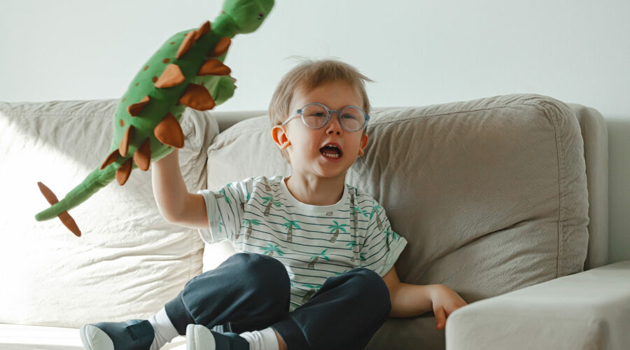 A child with autism in glasses sits on the sofa with his soft toy dinosaur.